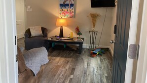 A living room with a sofa, a coffee table with books and a lamp, a painting on the wall, and a TV mounted above a door. - Available August 22-28! Cute and cozy (Ocala)