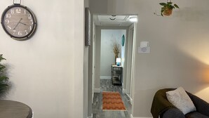 A hallway with a wooden floor, a round wall clock, a door leading to another room, and a small table with a lamp. - Available August 22-28! Cute and cozy (Ocala)