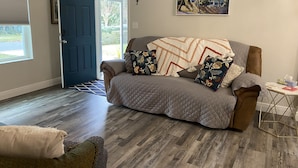 A cozy living room with a brown sofa, patterned pillows, and a quilted throw blanket. A painting hangs on the wall, and a small table with a tissue box is visible. - Available August 22-28! Cute and cozy (Ocala)