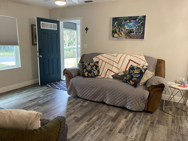 A cozy living room with a brown sofa, patterned pillows, and a quilted throw blanket. A painting hangs on the wall, and a small table with a tissue box is visible. - Crowd favorite! Clean and peaceful stay! (Ocala)
