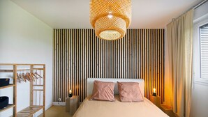 Un dormitorio con cama, lámpara colgante, pared de listones de madera, perchero con ganchos y ventana con cortinas.