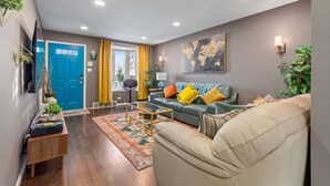 Een moderne woonkamer met een blauwe deur, een bank met gekleurde kussens, een bijzettafeltje en een groot abstract schilderij aan de muur.