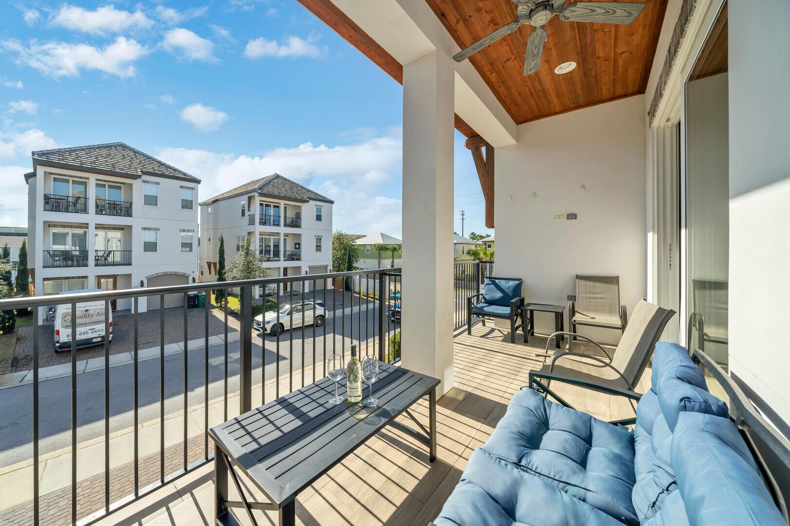 A balcony with a wooden deck, a blue cushioned sofa, a small table, and a chair. In the background, there are two multi-story buildings with balconies.