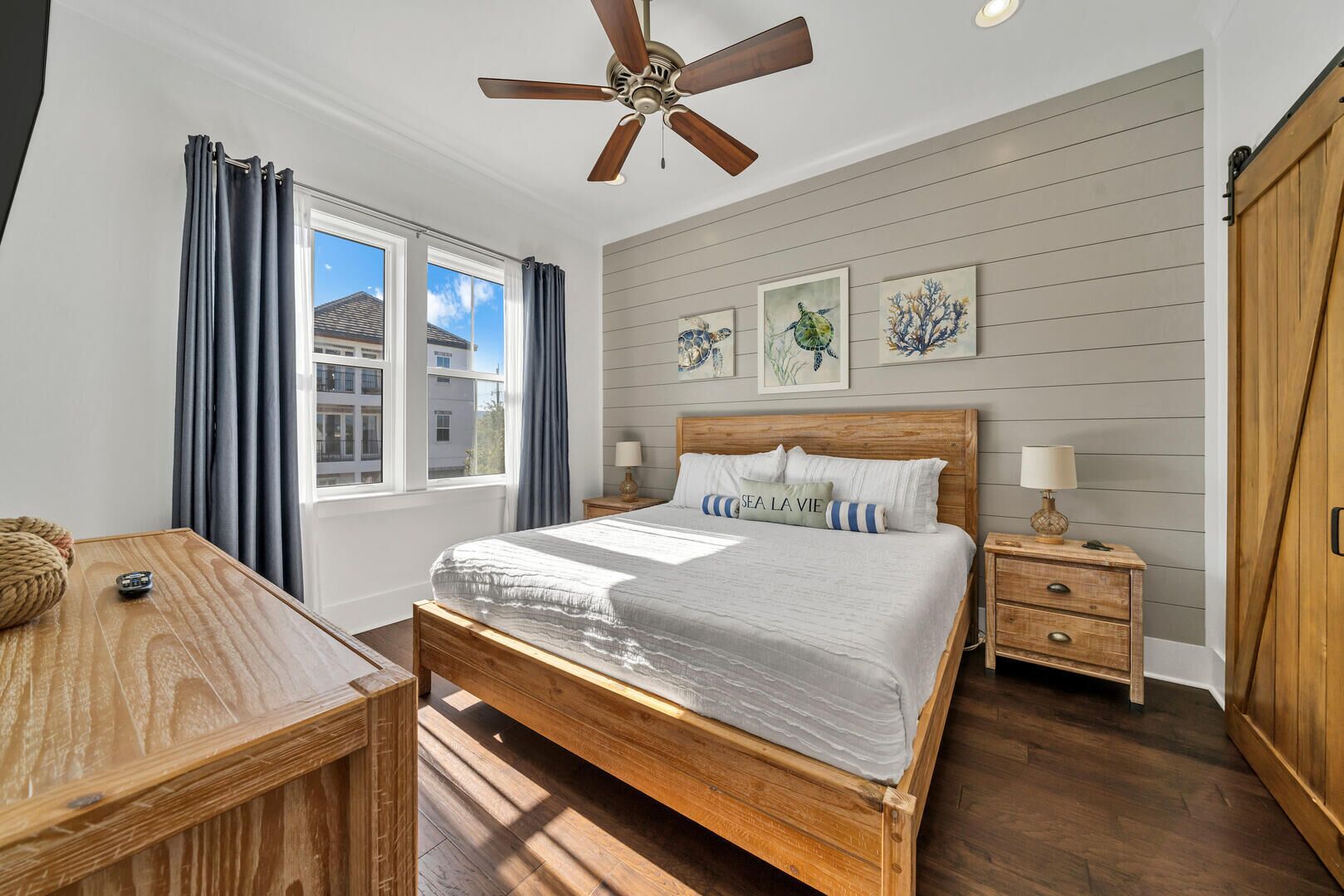 A bedroom with a wooden bed, a nightstand, a desk, a ceiling fan, and artwork on the wall.