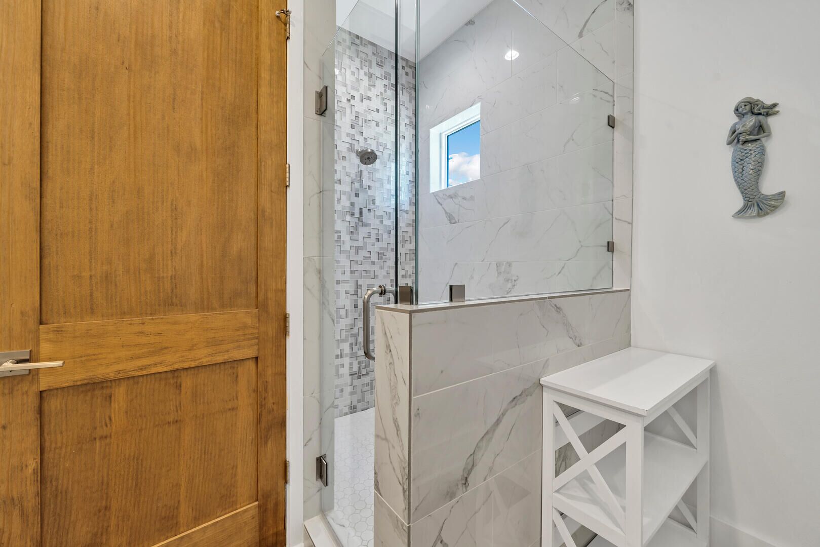 A bathroom with a wooden door, a glass shower enclosure, a marble-tiled shower area, and a white shelf.