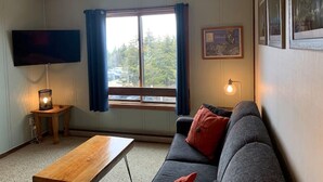 Una sala de estar con sofá, mesa de centro, televisor montado en la pared, ventana con cortinas y lámpara en una mesita auxiliar.