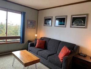 Un salon avec un canapé gris, deux coussins rouges, une table basse en bois et trois tableaux encadrés accrochés au mur.