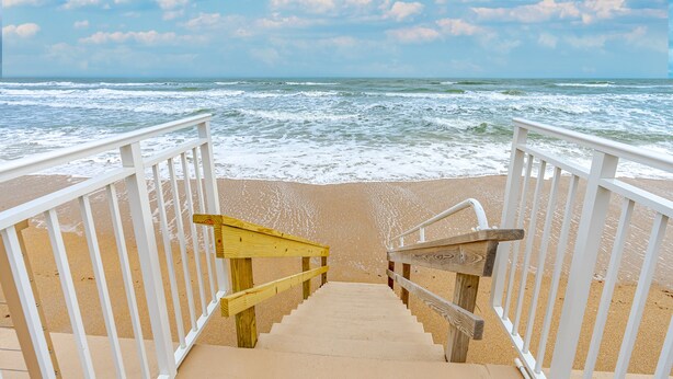 Una escalera de madera que lleva a una playa arenosa, con olas del océano al fondo.