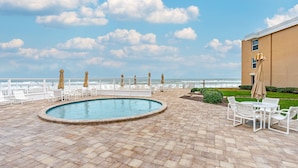 Un área de piscina con vista al océano, sillones blancos para tomar el sol y un edificio de fachada color beige.