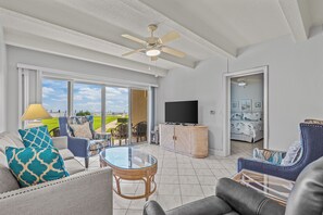 A spacious living room with a large window, a ceiling fan, and a flat-screen TV. - AMAZING OCEANVIEW - NO DRIVE BEACH - GROUND FLOOR - NEW SMYRNA BEACH FL (New Smyrna Beach)