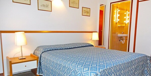 A hotel room with a bed, two bedside tables with lamps, a mirror with lights, and a door leading to a bathroom.