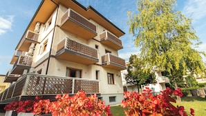 A multi-story residential building with balconies, a tree in the foreground, and red flowers in the foreground.