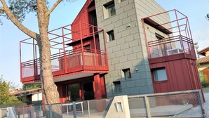 A modern building with a red and grey facade, multiple balconies with red railings, and a tree in the foreground.