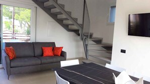 A modern living room with a grey sofa, red cushions, a dining table with chairs, a staircase with a metal railing, and a TV mounted on the wall.