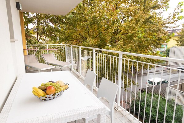 A balcony with a white table and chairs, a fruit bowl, and a view of a tree and street below.