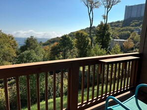 Un balcon donnant sur une colline boisée et sur un bâtiment situé au loin.