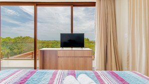 A hotel room with a bed, a TV on a wooden dresser, and a balcony with a view of trees and sky. - PF47 High Standard House 5 Suites - Praia do Forte (Mata de São João)