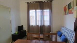 A living room with a sofa, a TV on a stand, a dining table with chairs, and a window with curtains.