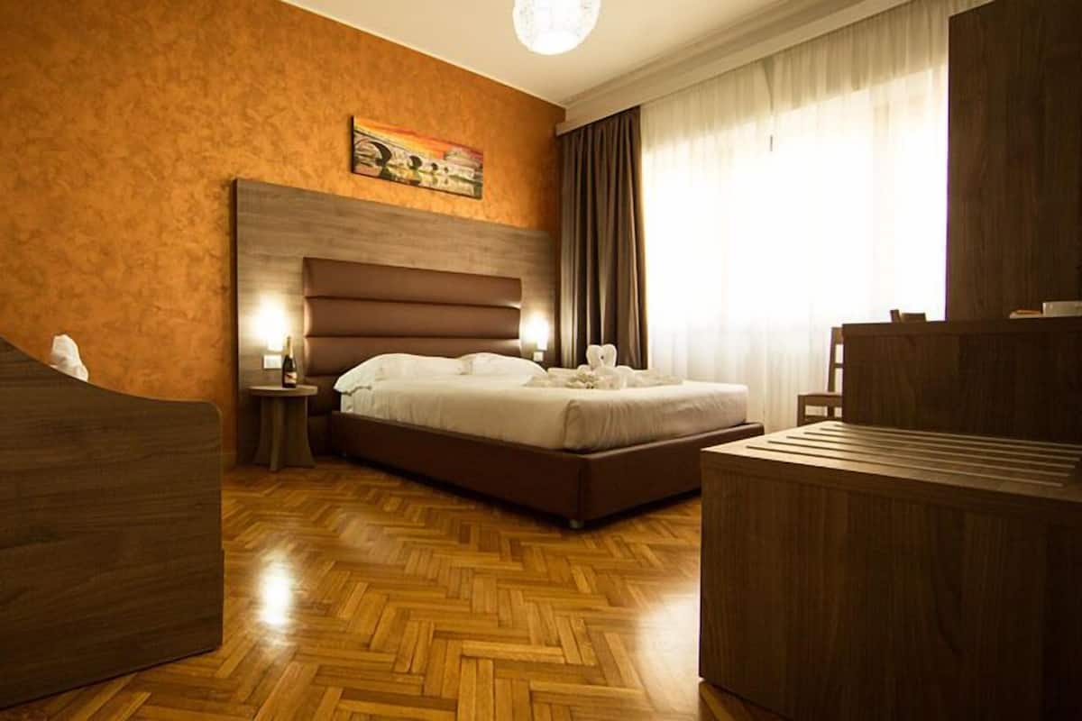 A hotel room with a large bed, a wooden headboard, a small table with a bottle, and a wooden desk.
