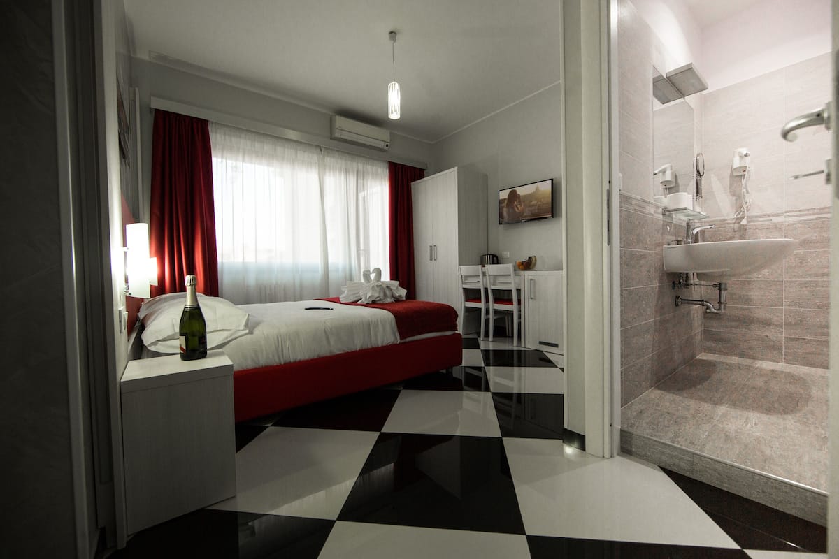 A modern hotel room with a checkered floor, a bed with red bedding, a bedside table with a bottle, a white wardrobe, a bathroom with a sink and mirror, and a shower area.