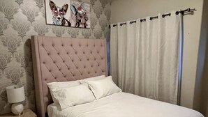 Un dormitori amb un panell de capçal acolxat, roba de llit blanca, una tauleta de nit amb una làmpada i una paret amb paper pintat estampat.
