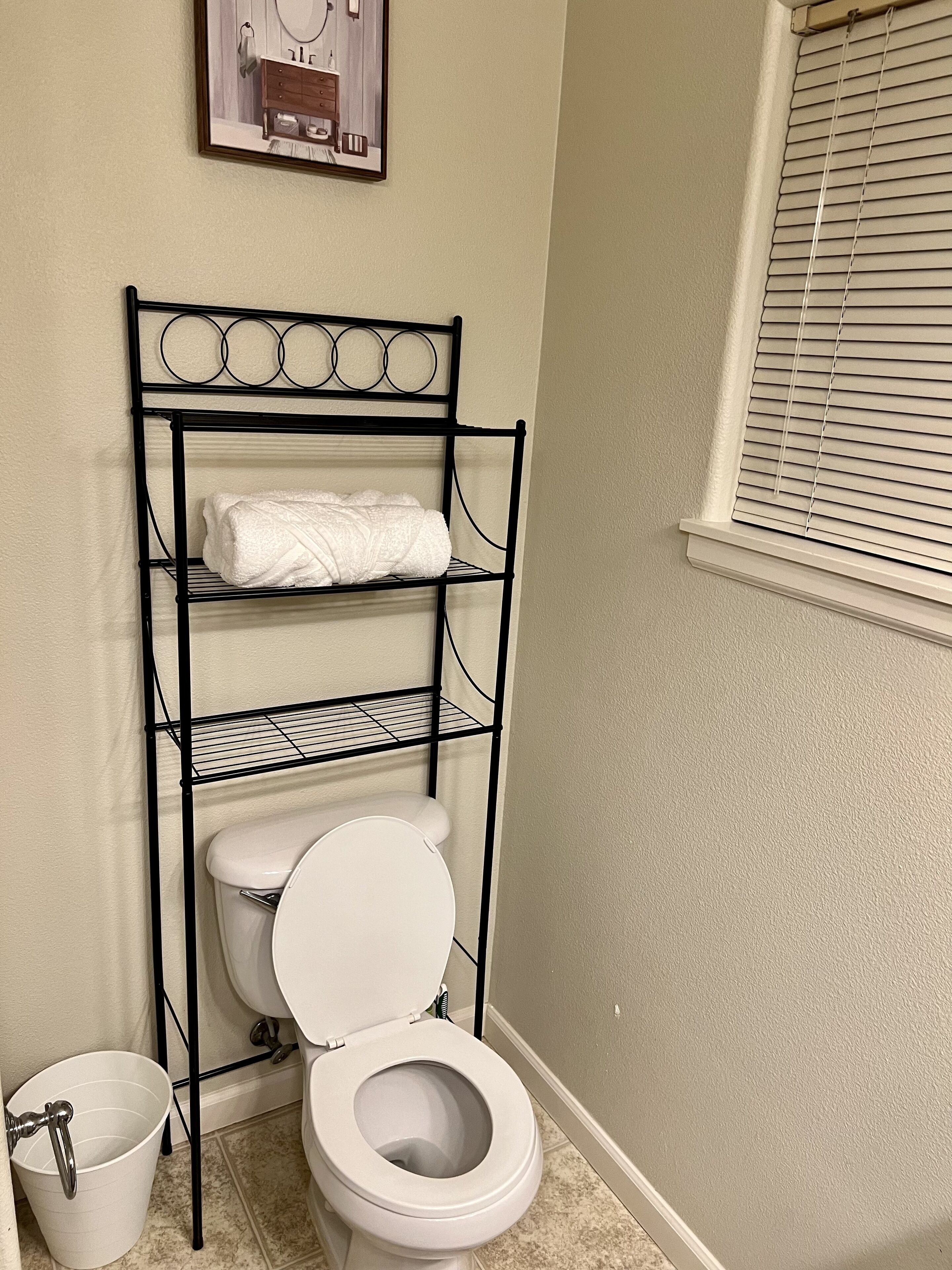 Un baño con un inodoro, una estantería metálica, un cubo blanco y una ventana con persianas.