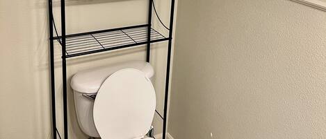 Un baño con un inodoro, una estantería metálica, un cubo blanco y una ventana con persianas.