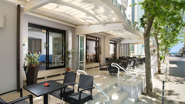 An outdoor seating area with tables and chairs, a glass partition, and a potted plant. - Hotel Mirabella (Cattolica)