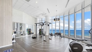Uma academia espaçosa com diversos equipamentos de exercício, grandes janelas com vista para o oceano e teto alto com luminárias modernas.