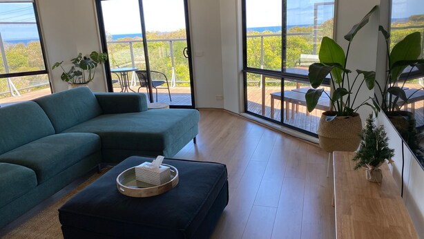 A modern living room with a sectional sofa, a coffee table, and large windows offering a view of the outdoors. - Beachfront at Cape Paterson (Cape Paterson)