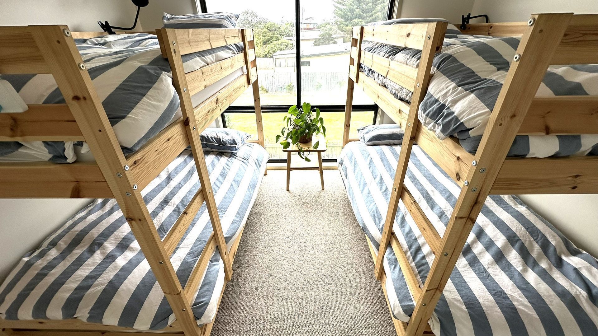 A room with three bunk beds, each with striped bedding, a small table with a plant, and a view of the outdoors.
