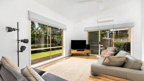 A spacious living room with a large TV, comfortable seating, and a view of the garden through sliding glass doors.