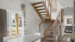 A modern interior with a wooden staircase, a dining area with white chairs, and a view of the outdoors.