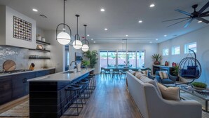 Una cocina moderna con una isla central, taburetes de bar e iluminación colgante. Hay un comedor con un sofá y sillas, además de un ventilador de techo.