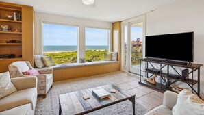 A living room with a large TV, a bookshelf, a window seat with a view of the ocean, and a ceiling fan.