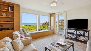 A living room with a large TV, a sofa, a coffee table, and a bookshelf. There are large windows with a view of the ocean.