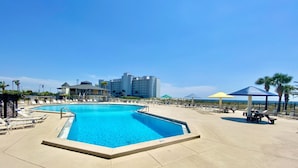 Una piscina con lettini e ombrelloni, circondata da edifici e palme.