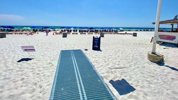 Una spiaggia con un tappetino blu che conduce a un'area di ritiro sedie, un cartello per il noleggio di paddleboard e un cartello che indica un costo per il parcheggio oltre la spiaggia.