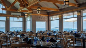 Un restaurant avec des tables et des chaises en bois, de grandes fenêtres donnant sur l’océan et des lampes suspendues de style lanterne.