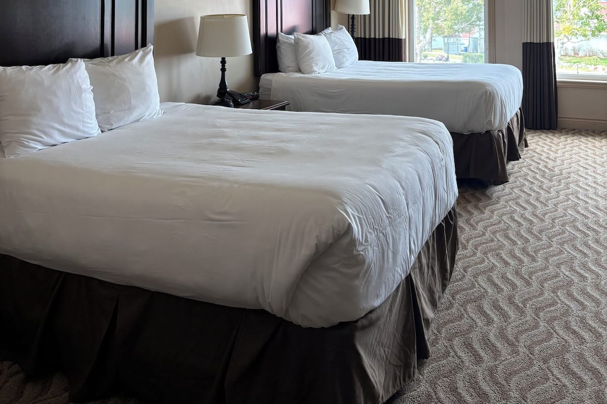 A hotel room with two beds, a dark wood headboard, a large window with curtains, and a framed picture on the wall.