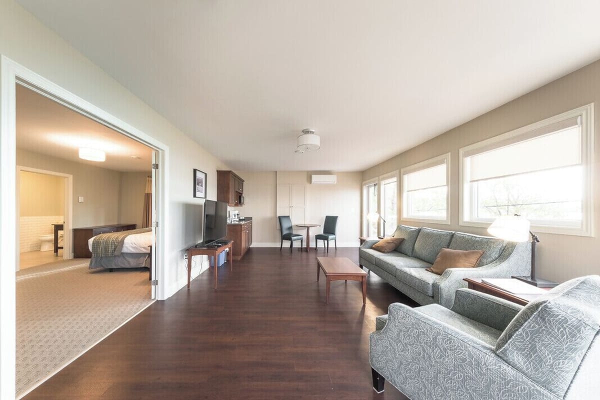A spacious living area with a sofa, a coffee table, and a dining table. There is a kitchen area with cabinets and a refrigerator. The room has large windows allowing natural light.