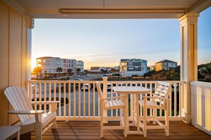 A wooden deck with two Adirondack chairs and a small table, overlooking a street with buildings and a sunset. - Salty Girl: Coastal Condo Close to Pier, Restaurants (Oak Island)
