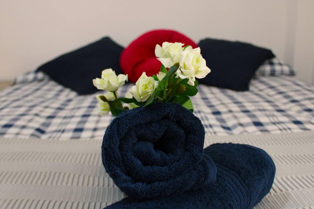 Ein ordentlich bezogenes Bett mit einer karierten Tagesdecke, ein aufgerolltes blaues Handtuch, weiße Blumen und zwei Kissen, eines rot und eines schwarz.