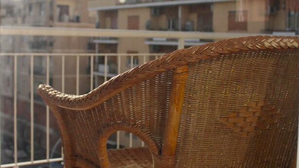 Une chaise en osier sur un balcon, avec en arrière-plan la vue sur des immeubles.