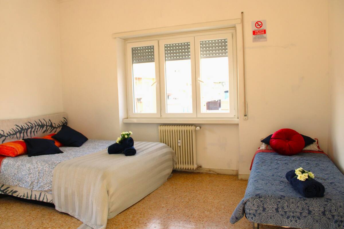 Ein Zimmer mit zwei Betten, einem Fenster mit Jalousien, einem Heizkörper und einem Rauchverbotsschild an der Wand.