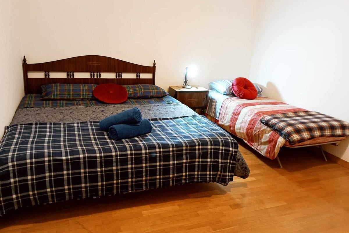 Ein Zimmer mit zwei Betten, einem größeren und einem kleineren, beide mit Karomuster-Bettwäsche. Dazwischen steht ein Nachttisch mit einer Lampe.