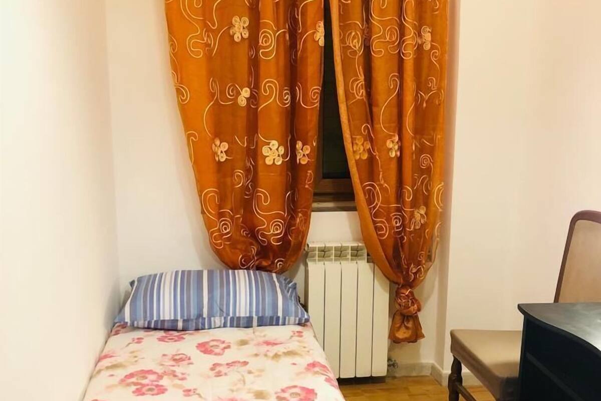 Ein kleines Schlafzimmer mit Bett, Heizkörper, Schreibtisch und orangefarbenen Vorhängen.
