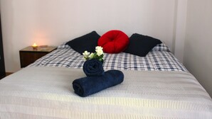 Un lit bien fait, recouvert d’une couverture à carreaux, avec une serviette bleu foncé enroulée, un oreiller rouge et un petit bouquet de fleurs blanches.