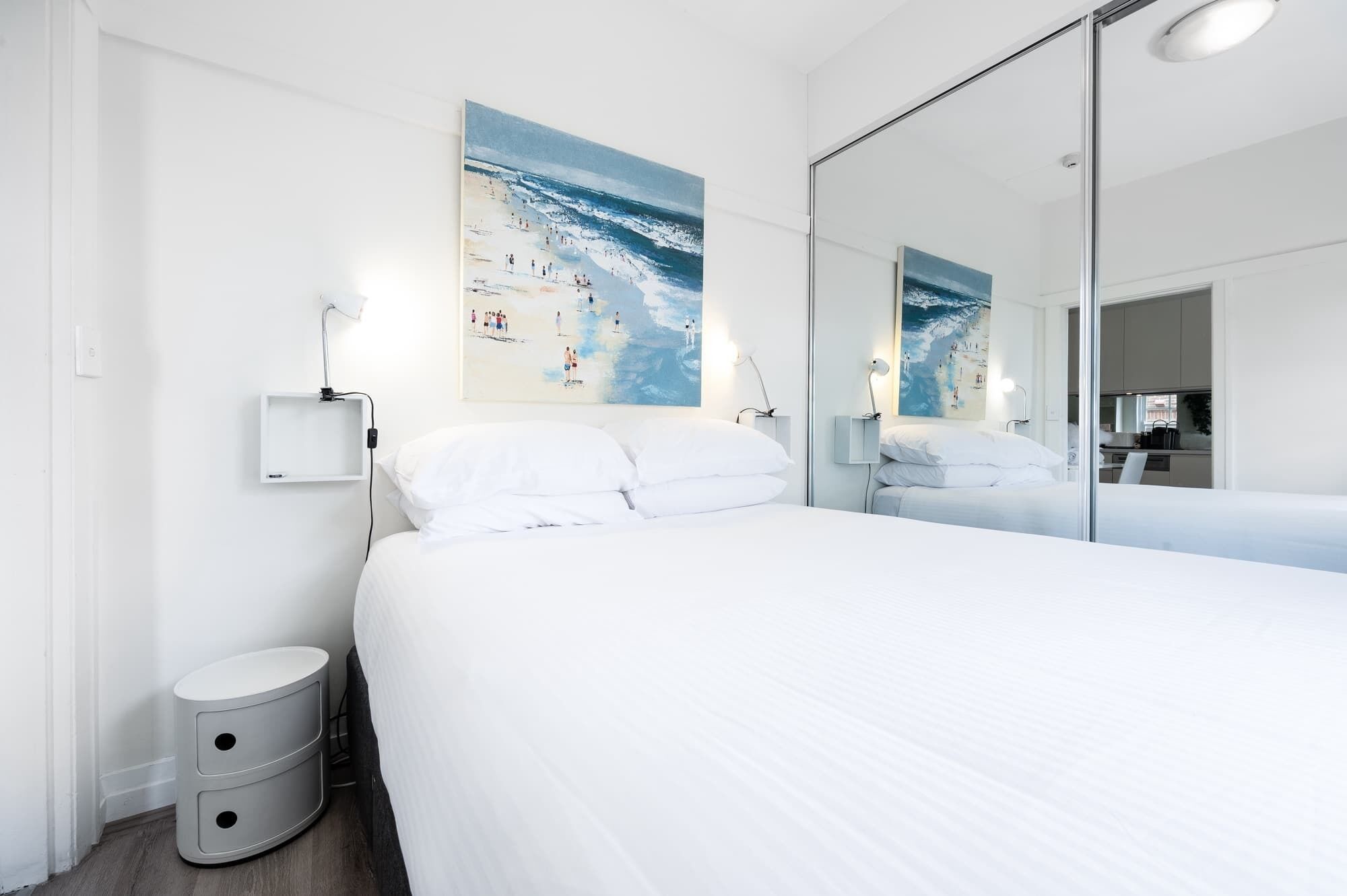 Ett modernt hotellrum med en stor säng, ett nattduksbord, en liten papperskorg och en stor spegel som reflekterar rummet.
