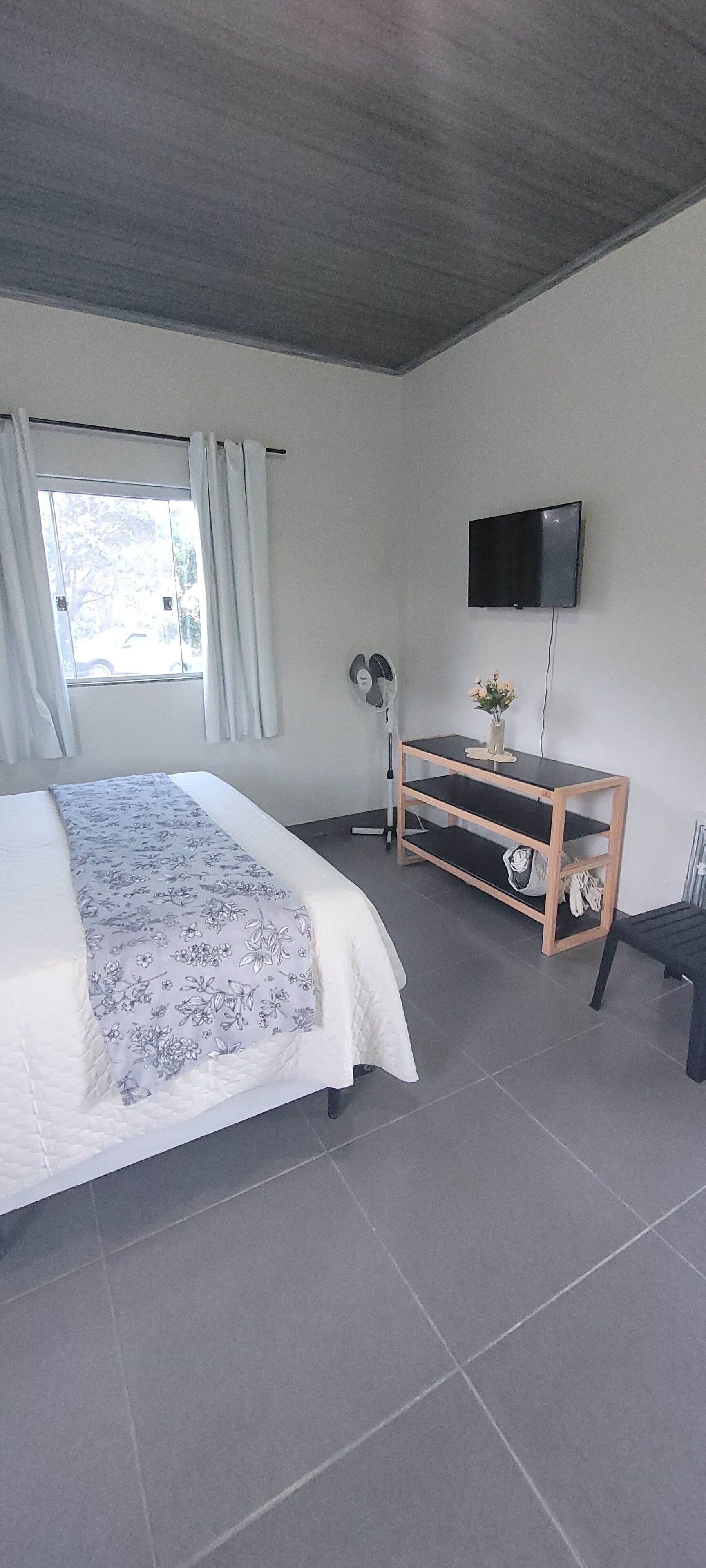 A bedroom with a bed, a TV mounted on the wall, a wooden side table, and a window with curtains.